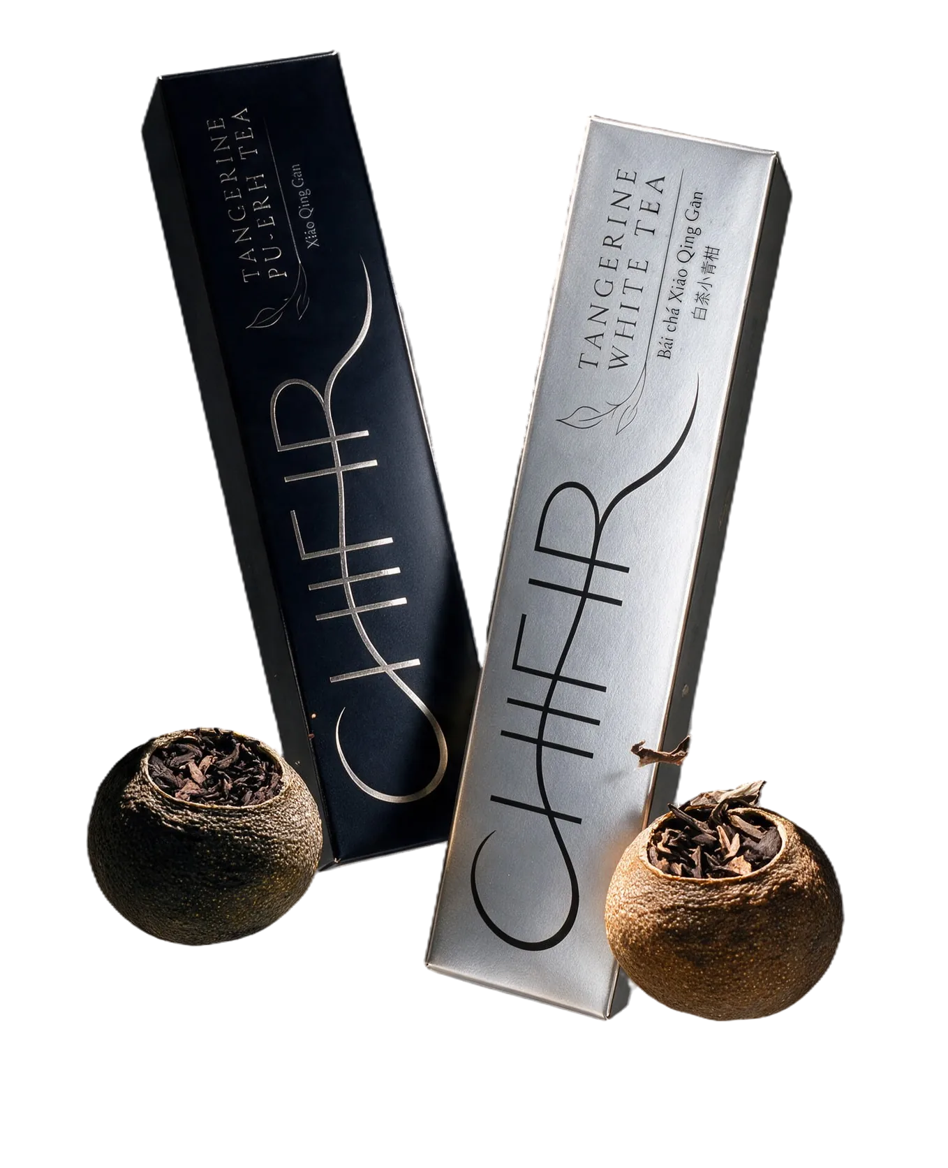 Two CHIFIR cartons floating at an angle — matte black Tangerine Pu-erh and silver Tangerine White — among opened sun-dried green tangerines, their cut lids, dried leaves and tea crumbs, casting a soft shadow on a seamless pale grey.