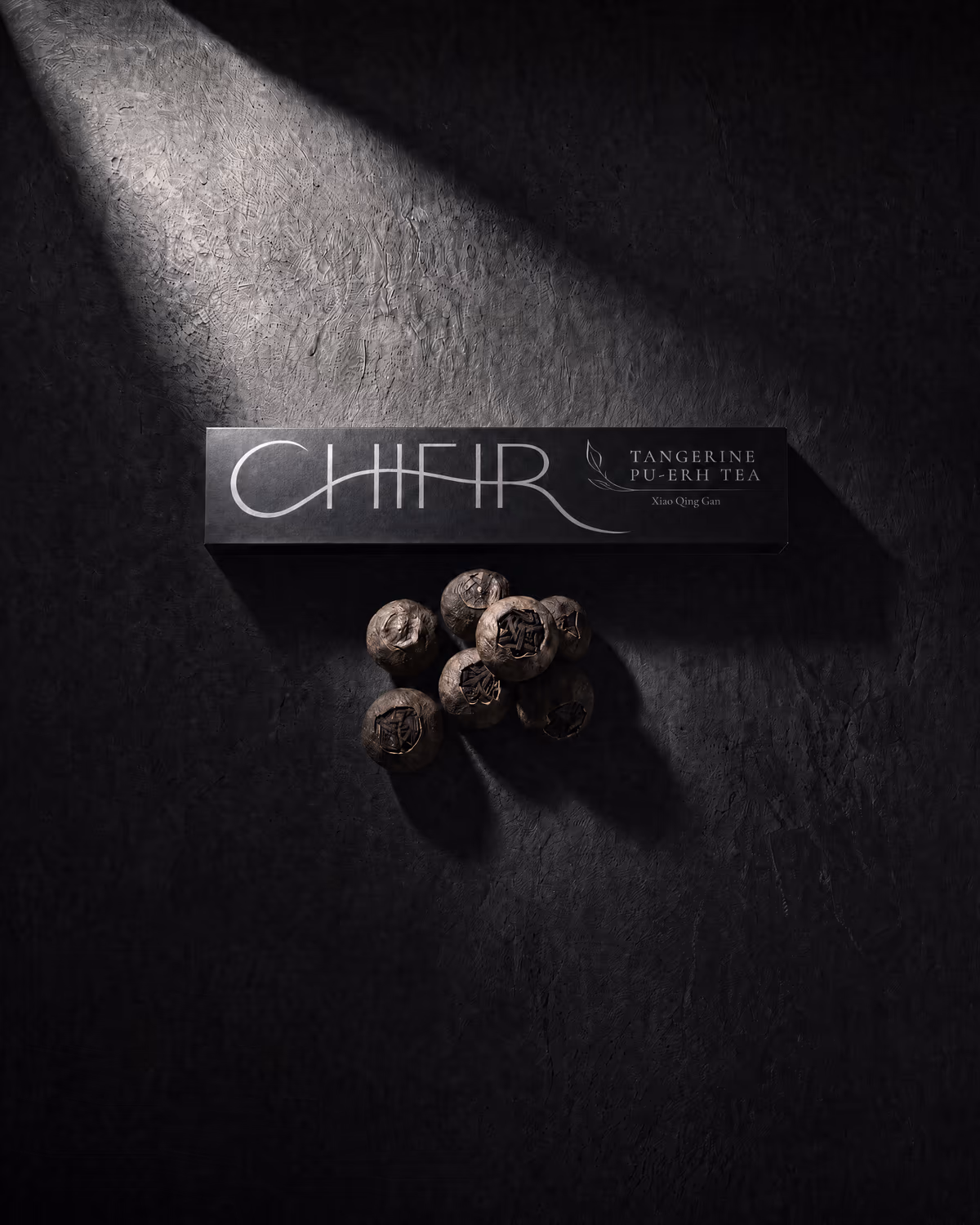 The black box — Tangerine Pu-erh Tea carton on dark stone, a cluster of leaf-stuffed tangerines beside it under a slant of light.