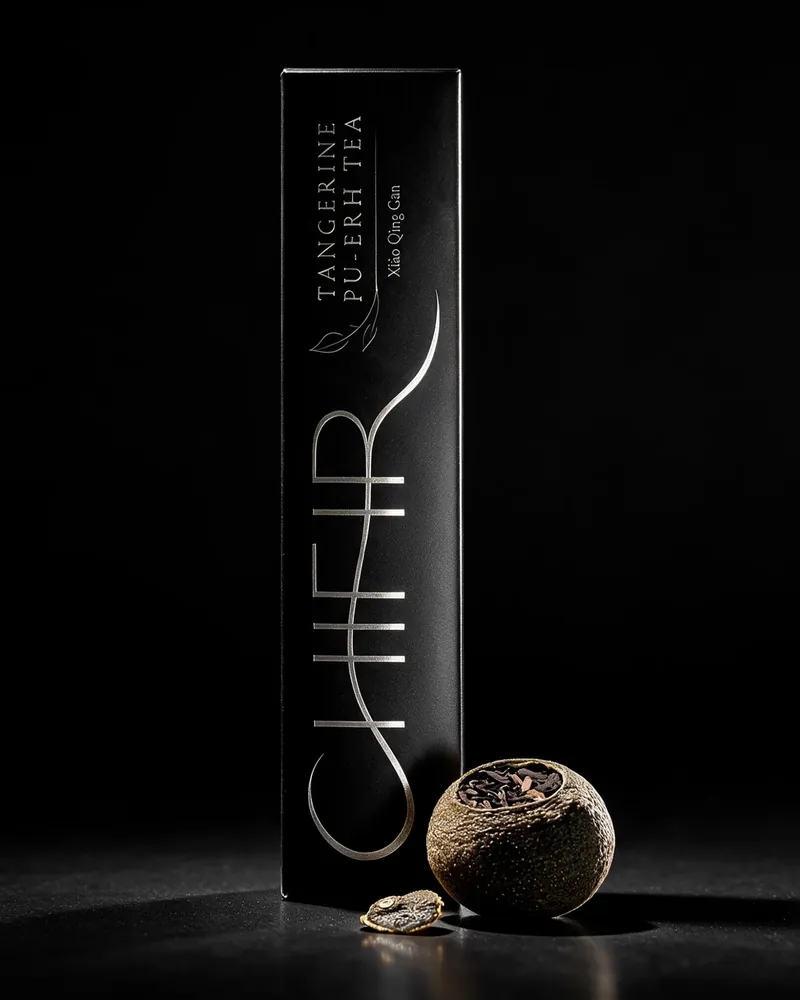 The black carton standing upright on charcoal, one whole dried pu-erh tangerine and its cut stem cap on the stone beside it.