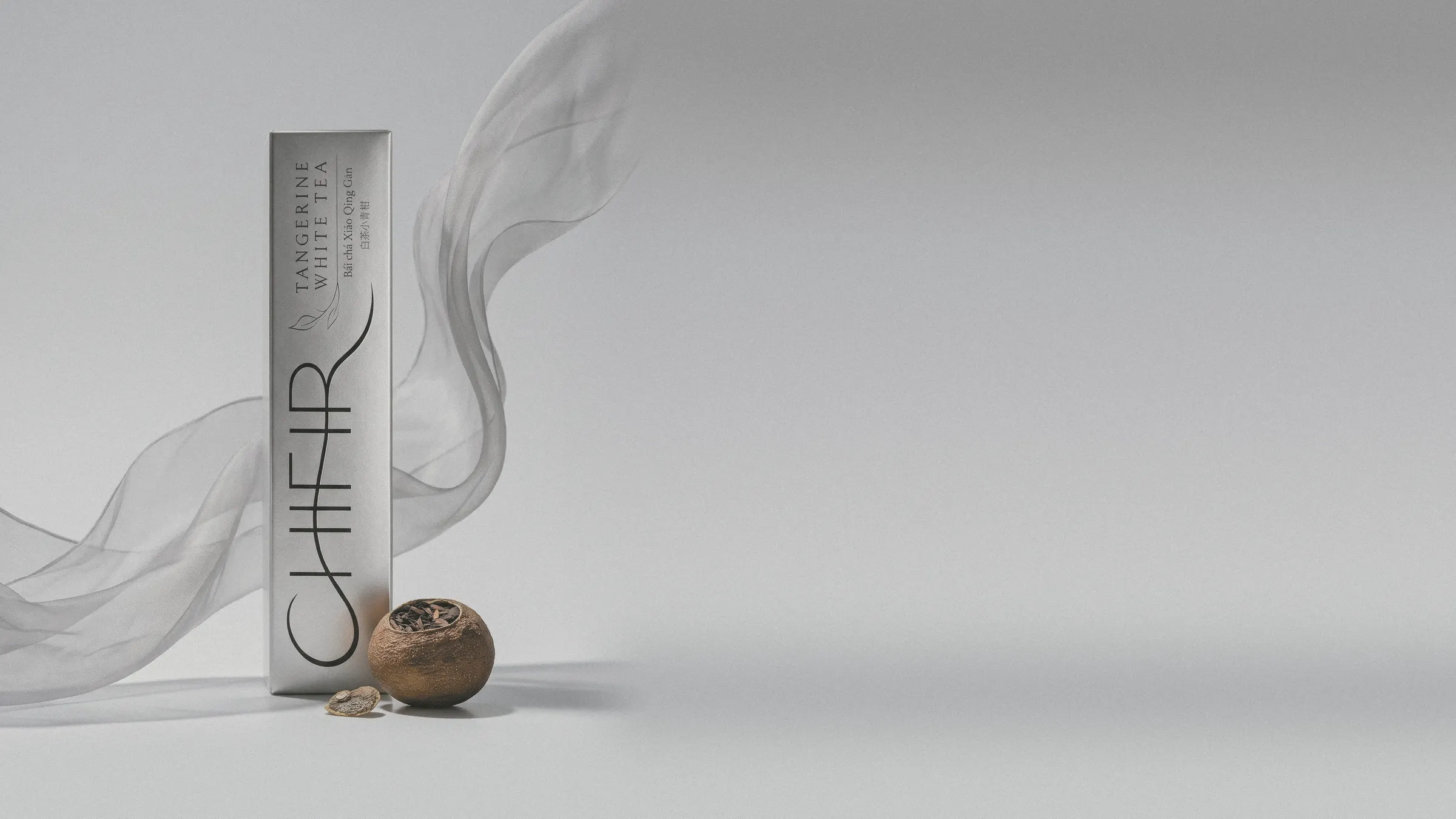 The silver Tangerine White carton at the left in pale studio light, a sheer veil of silk organza curving past it, a dried tangerine piece at its foot; the pale ground runs on to the right.
