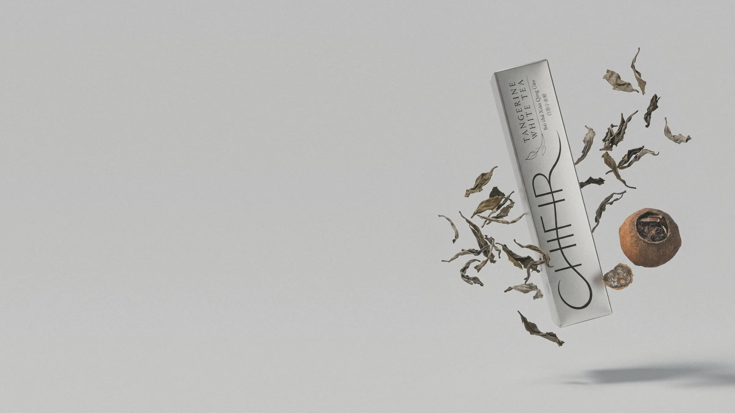 The silver carton floating at an angle at the right, dried white tea leaves and a tangerine piece drifting around it, on a pale studio ground that runs on to the left.