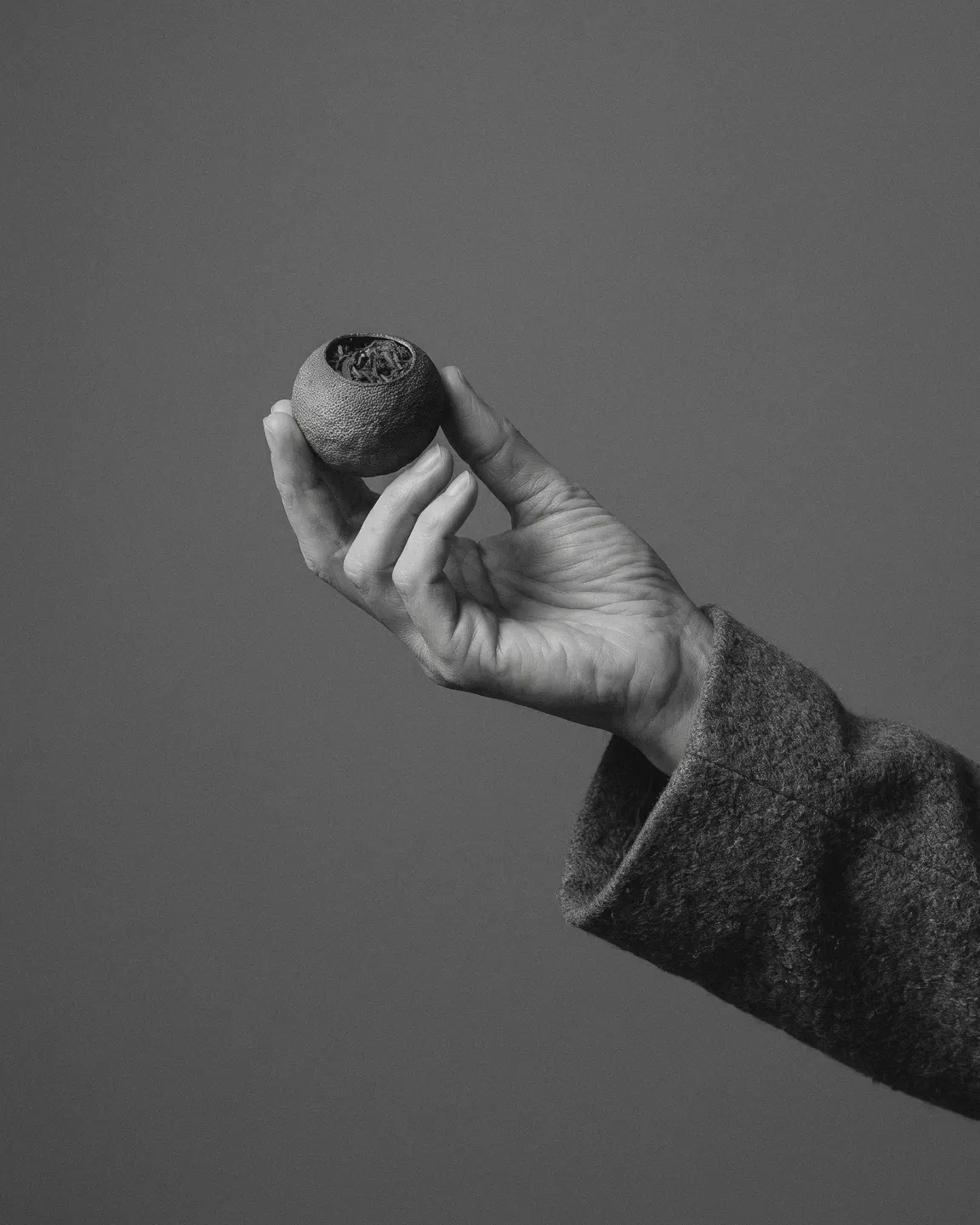 A hand in a charcoal wool sleeve, palm open, holding one dried tangerine piece against deep shadow.