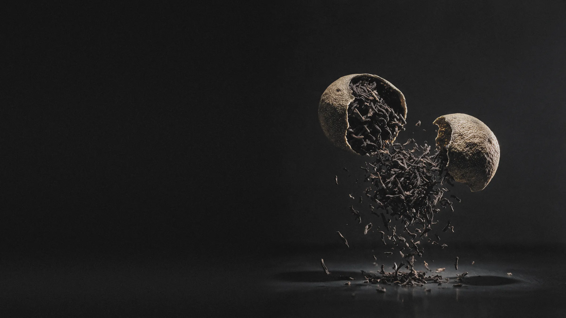 A dried tangerine piece cracked open in mid-air at the right, ripe pu-erh leaf falling out of it onto a charcoal studio ground that runs on to the left.