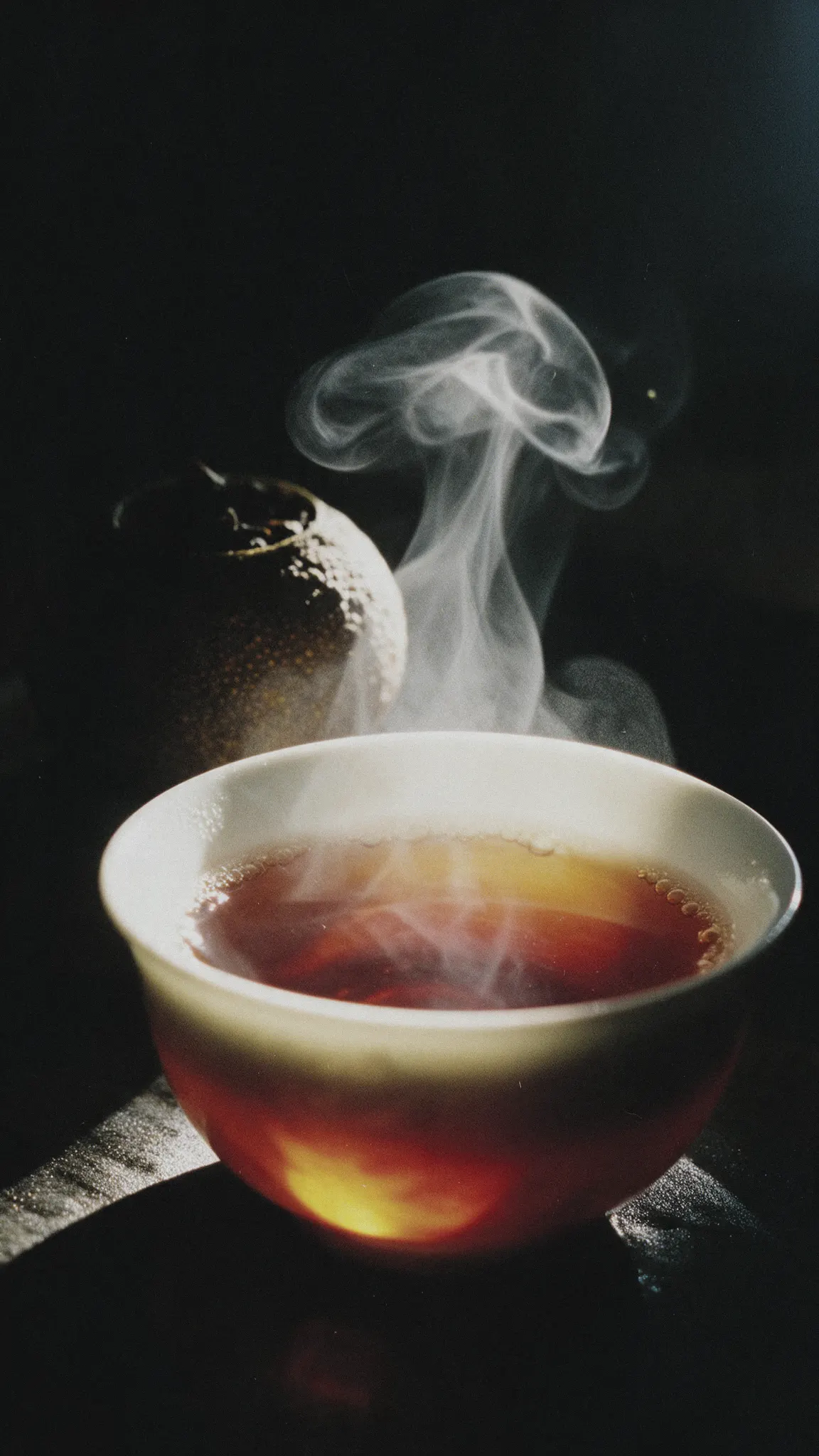 A small white cup of red-amber Tangerine Pu-erh, steam rising from it, one whole dried tangerine piece behind it in the dark.