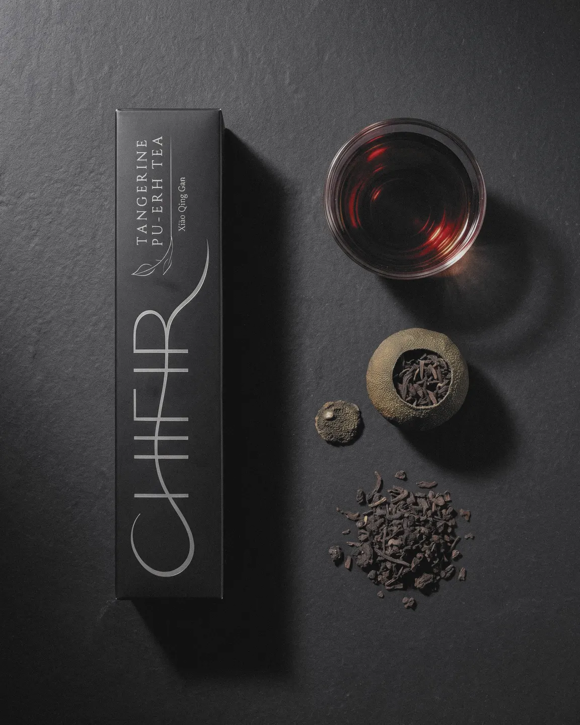 From above, on charcoal: the black carton, a glass cup of dark amber tea, one tangerine piece with its cap, and a small heap of loose ripe pu-erh leaf.