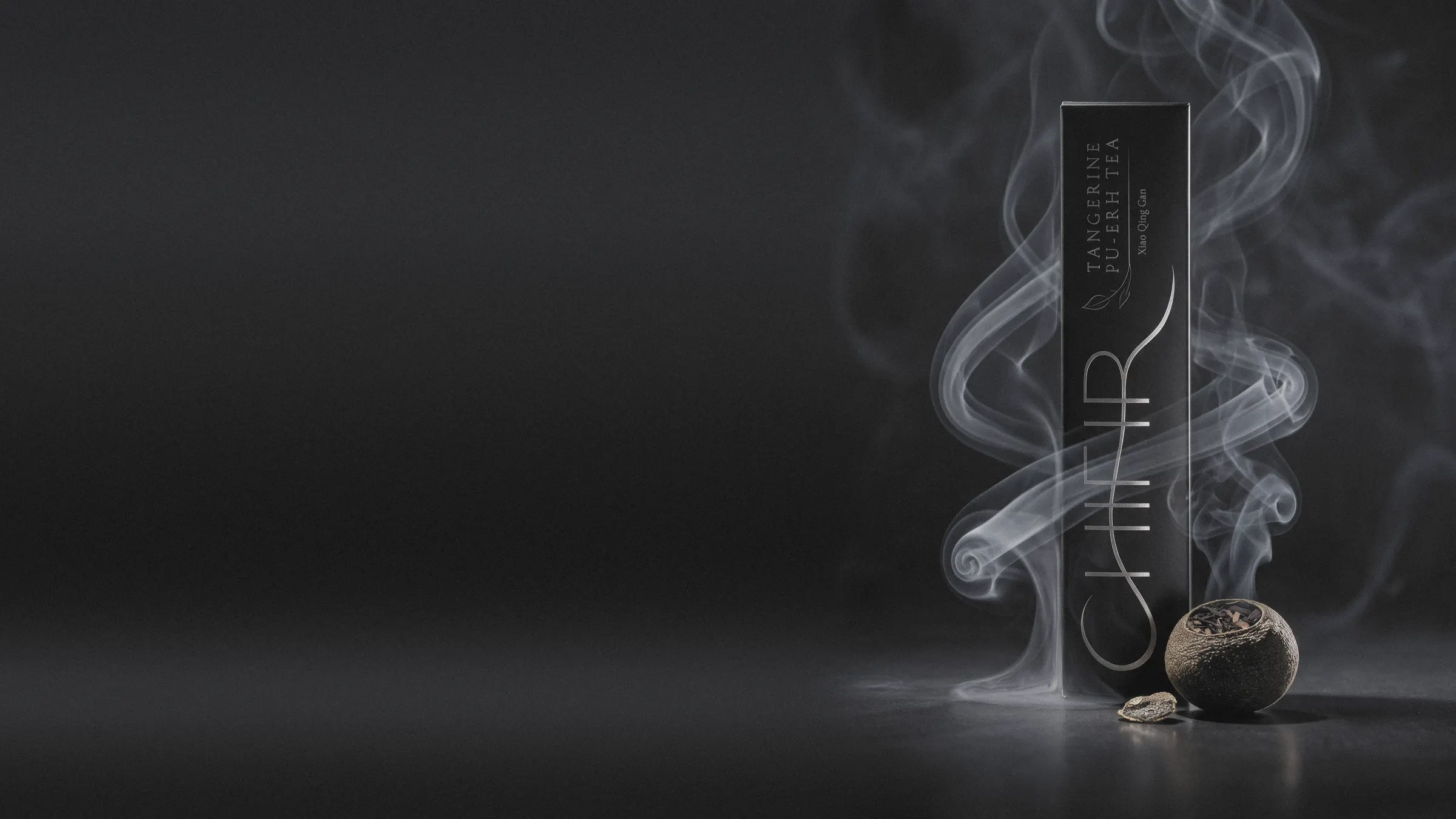 The black Tangerine Pu-erh carton standing in near-darkness at the right, a slow ribbon of pale smoke curling around it, one dried tangerine piece at its foot; the charcoal studio ground runs on to the left.