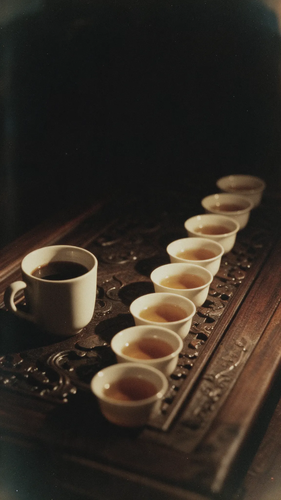 Eight small cups of amber tea in a diagonal row on a carved wooden tray, one full coffee mug beside them.